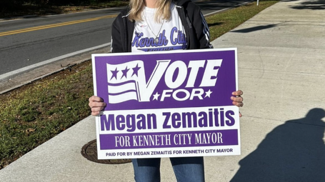 A woman in a cap holding a sign to vote for her in Kenneth City