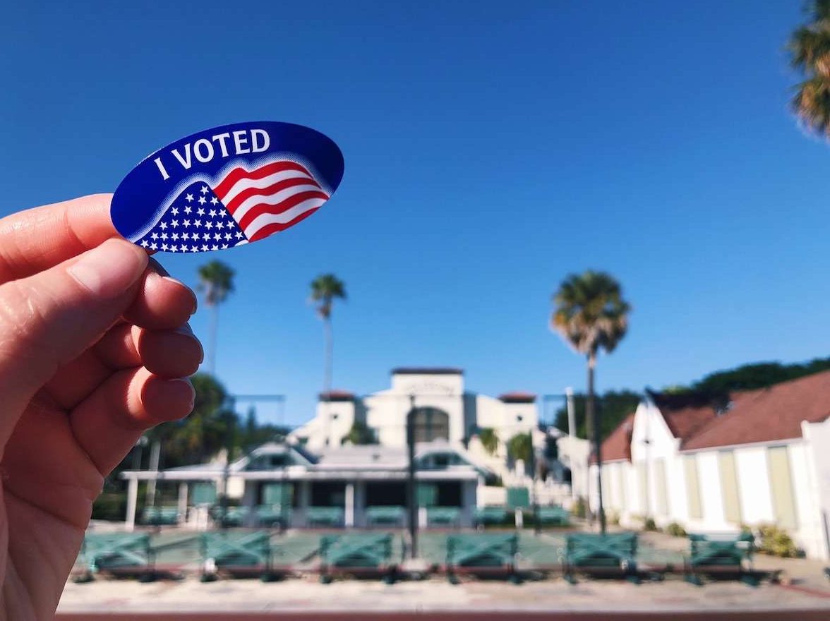 a person's hand holding an "i voted" sticker in front of the st. petersburg shuffleboard club. (2025 South Pinellas Municipal Election Candidates)