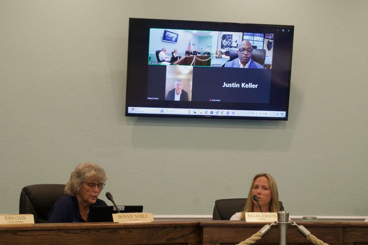 TV in background shows four squares. One of them is black. Closer, Megan Zemaitis sits on the right and Bonnie Noble sits on the left behind a brown desk.