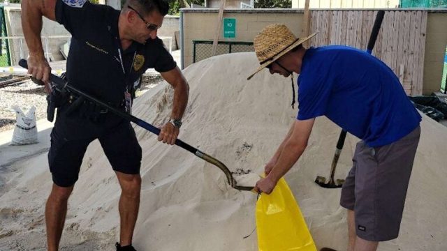 a cop shoveling sand into a yellow bag that a man holds open.