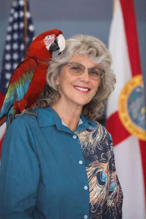 woman with a parrot on her shoulder sitting in front of an American flag.