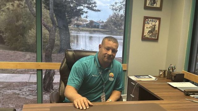 a man in a polo shirt sitting at a big wooden desk.