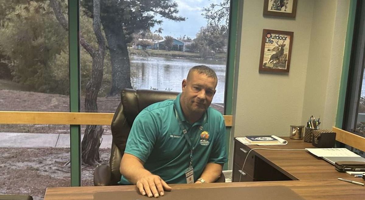a man in a polo shirt sitting at a big wooden desk.