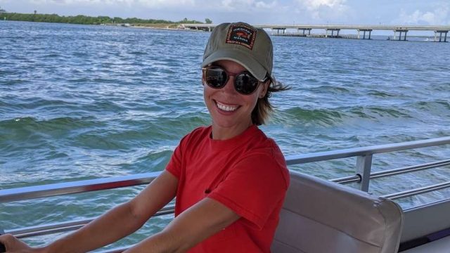 A woman with a orange shirt, sunglasses, and a hat driving a boat.