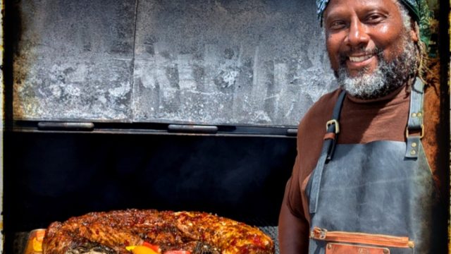 a man smiling while next to a grill of him with cooked meat.