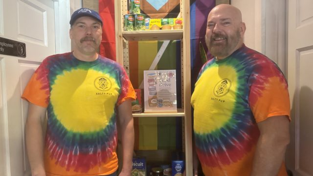 Two men wearing tie-dye shirts standing in front of a shelf.