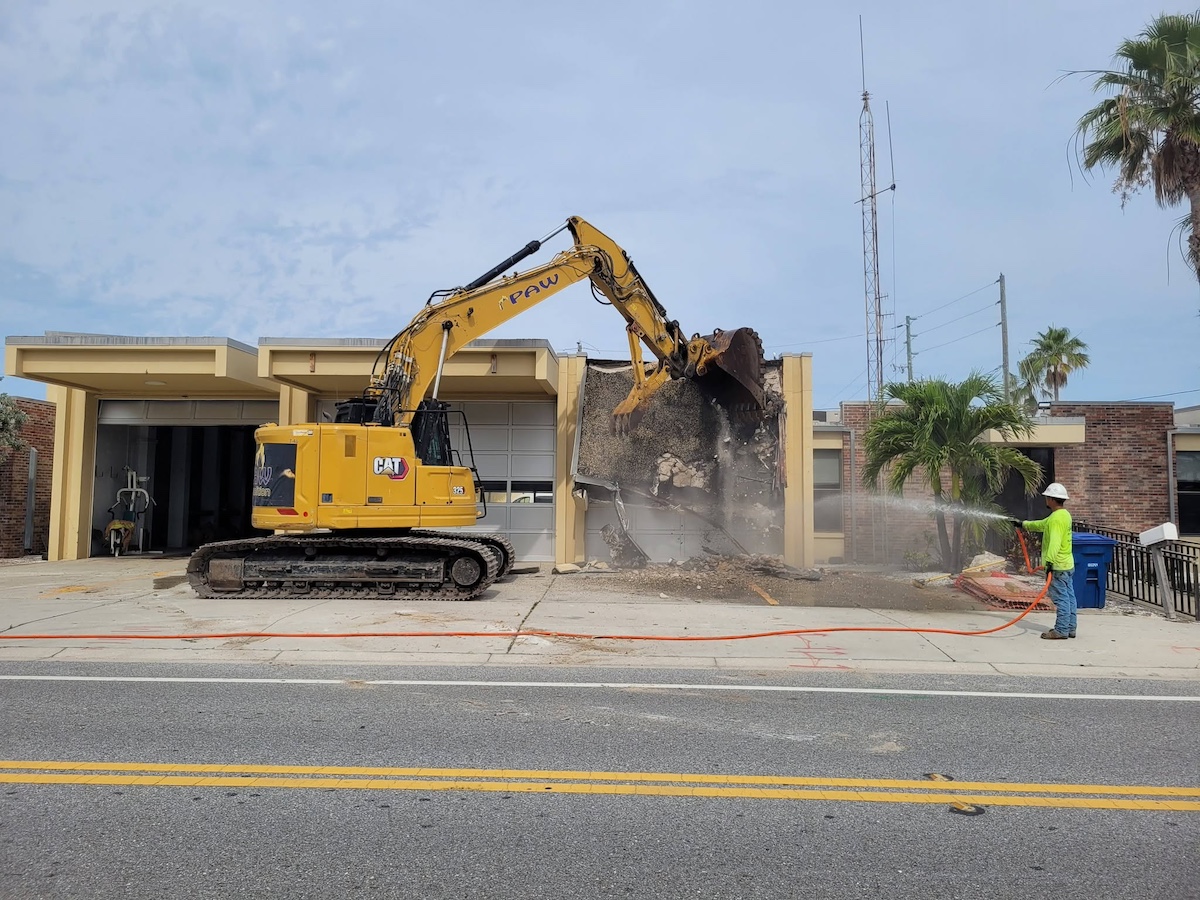 An excavator in front of a building and a person using a hose.