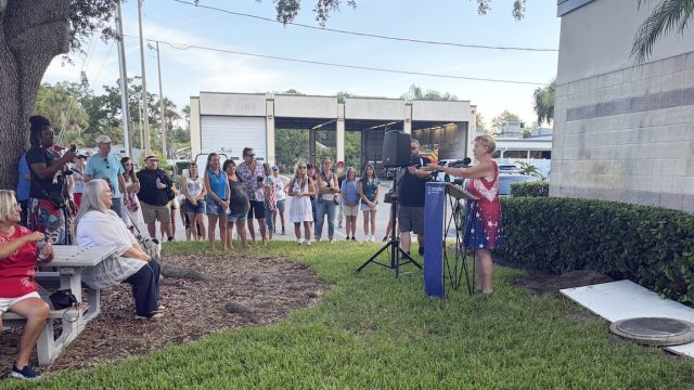 people standing outside and a woman speaking into a mic.