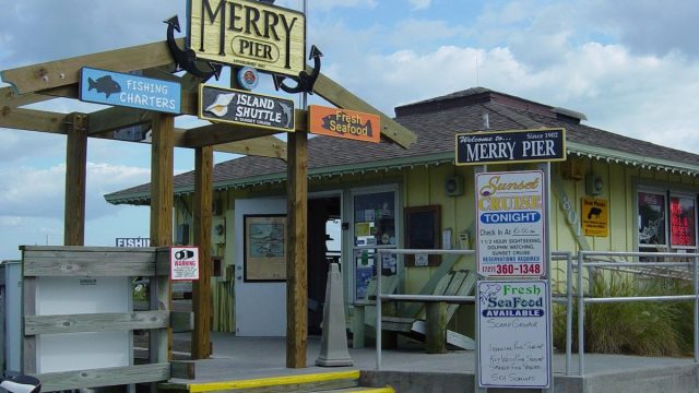 The entrance to Merry Pier in Pass-a-Grille, with signs advertising fishing charters, an island shuttle, fresh seafood and sunset cruises.