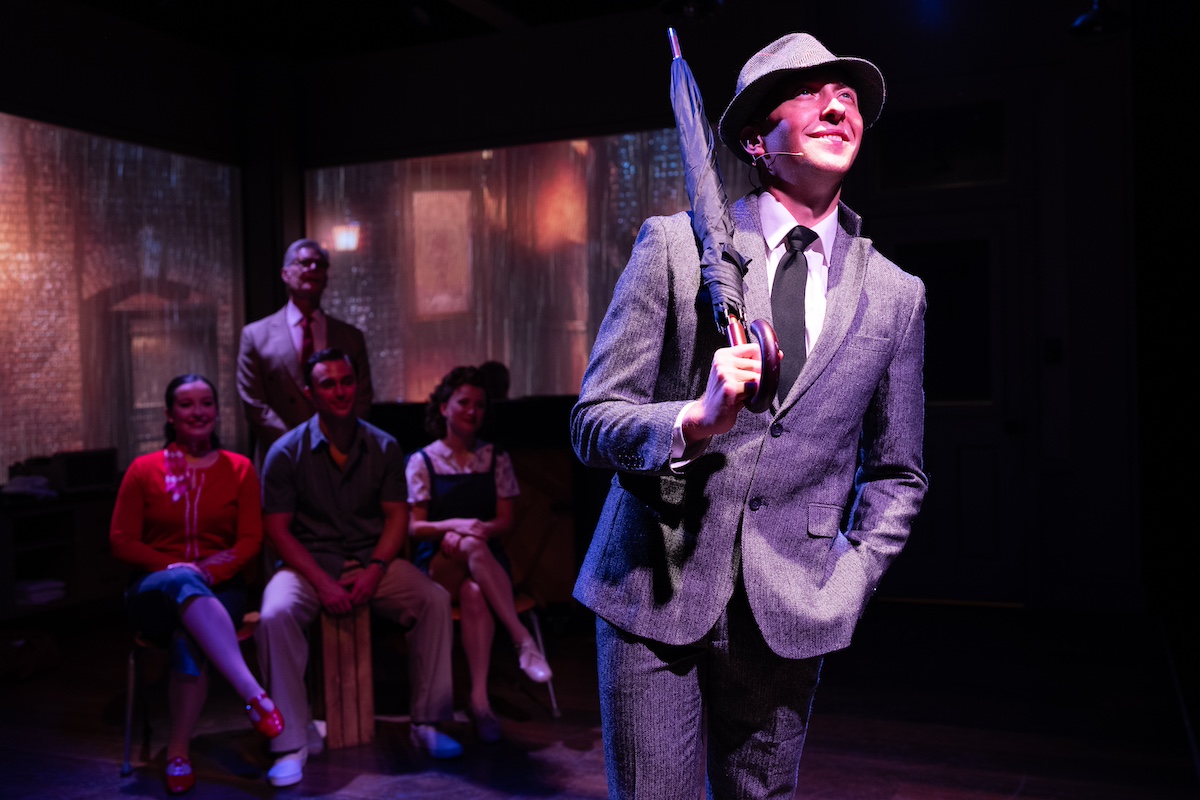 a man in a hat and suit holding a folded umbrella in front of four people sitting behind him.
